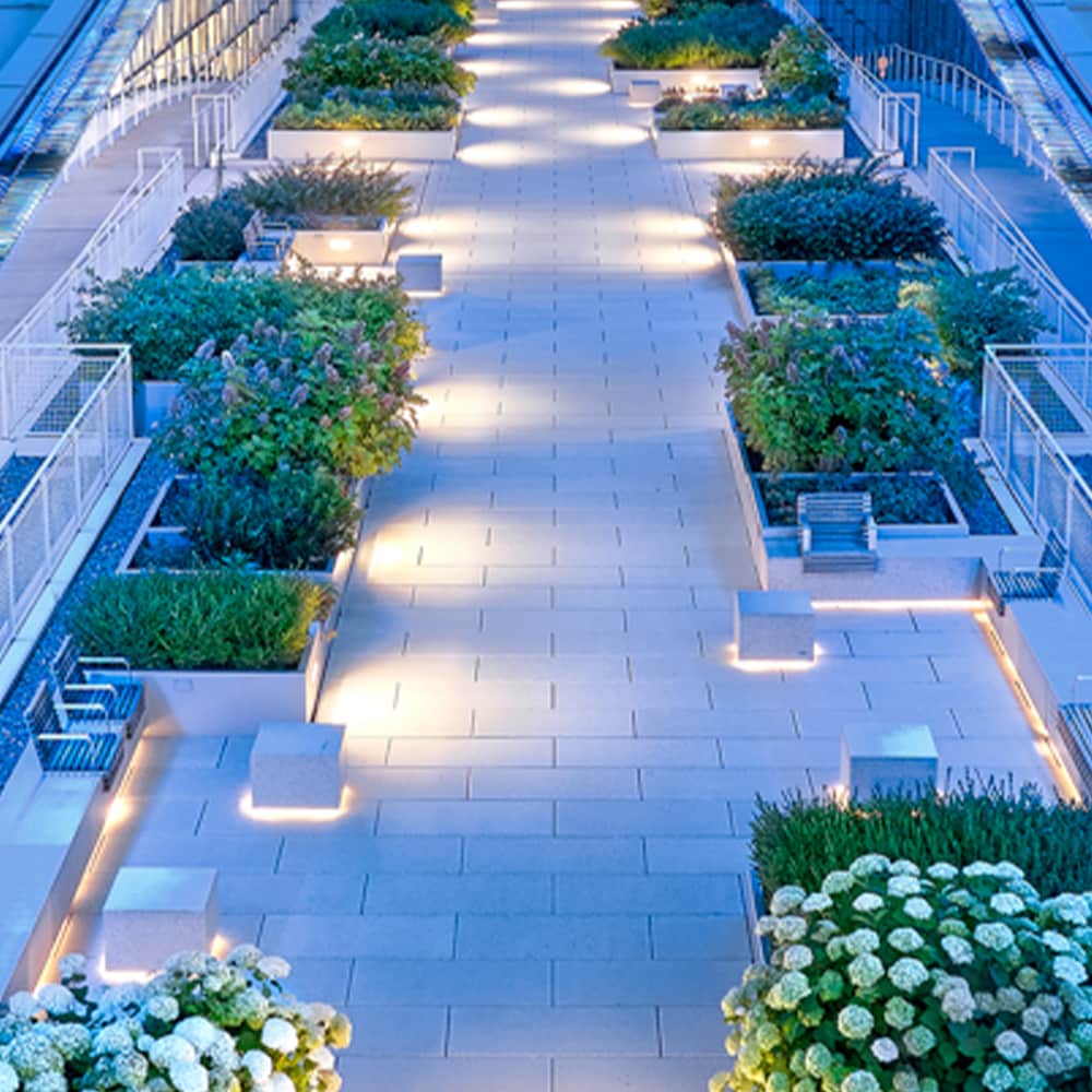 Nighttime overhead view of an illuminated commercial green roof plaza with pavers and landscaped planters featured on the Siplast RISE blog.