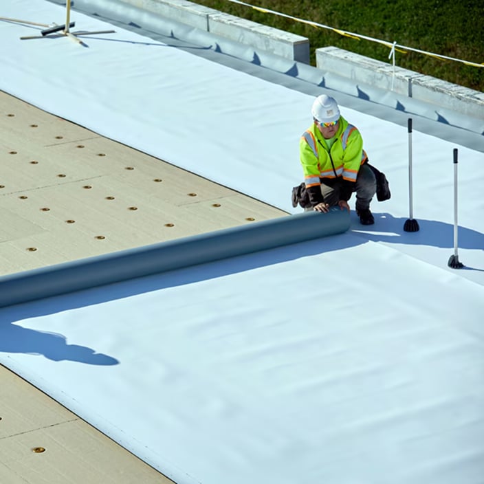 Roofing contractor unrolling a Siplast Parasolo single-ply membrane over cover board on a commercial flat roof.