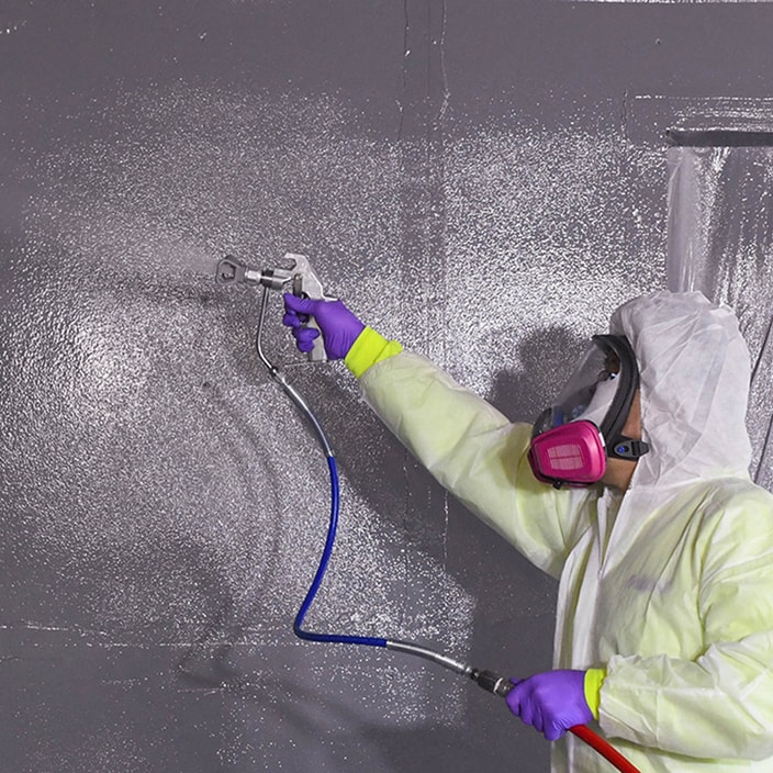 Professional applicator in protective suit spray-applying Siplast WALLcontrol fluid-applied air and water-resistive wall barrier.