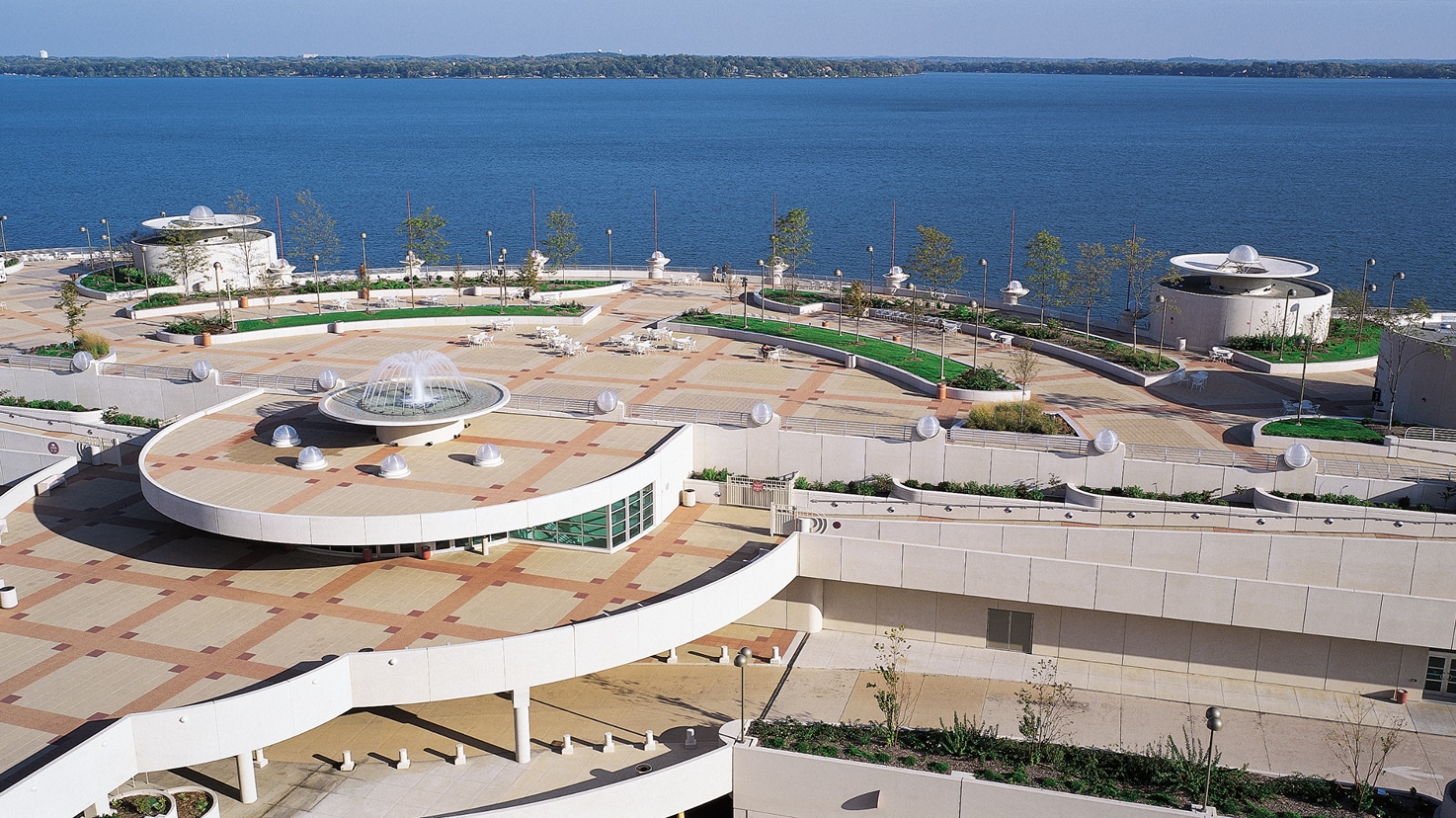 Elevated outdoor space featuring geometric paving, circular structures, and fountain, overlooking a lake