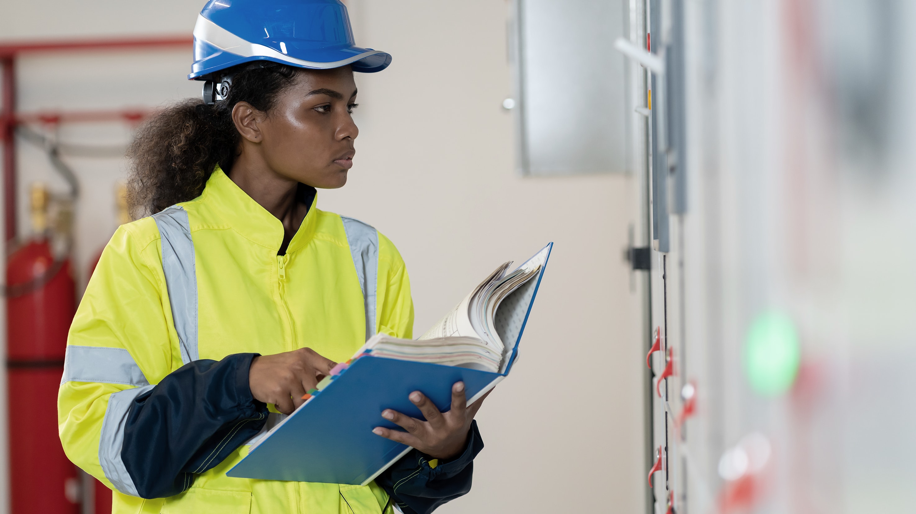 Construction worker in safety gear holding a tablet and clipboard, inspecting equipment at a construction site with visible piping and machinery