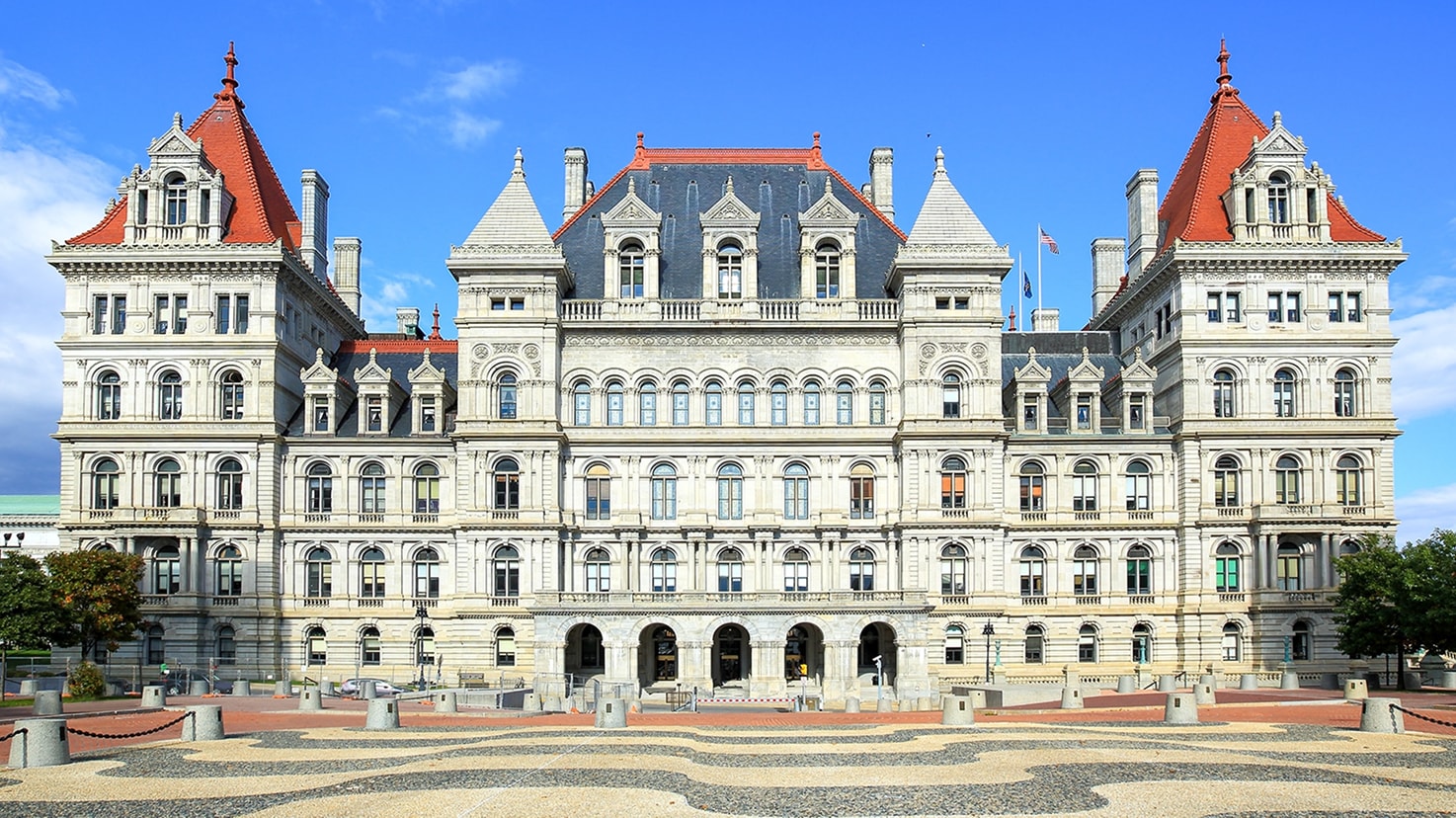 Exterior facade of the New York State Capitol building in Albany, New York, featuring the historic stone structure, arched entrances, and prominent red slate roofs.