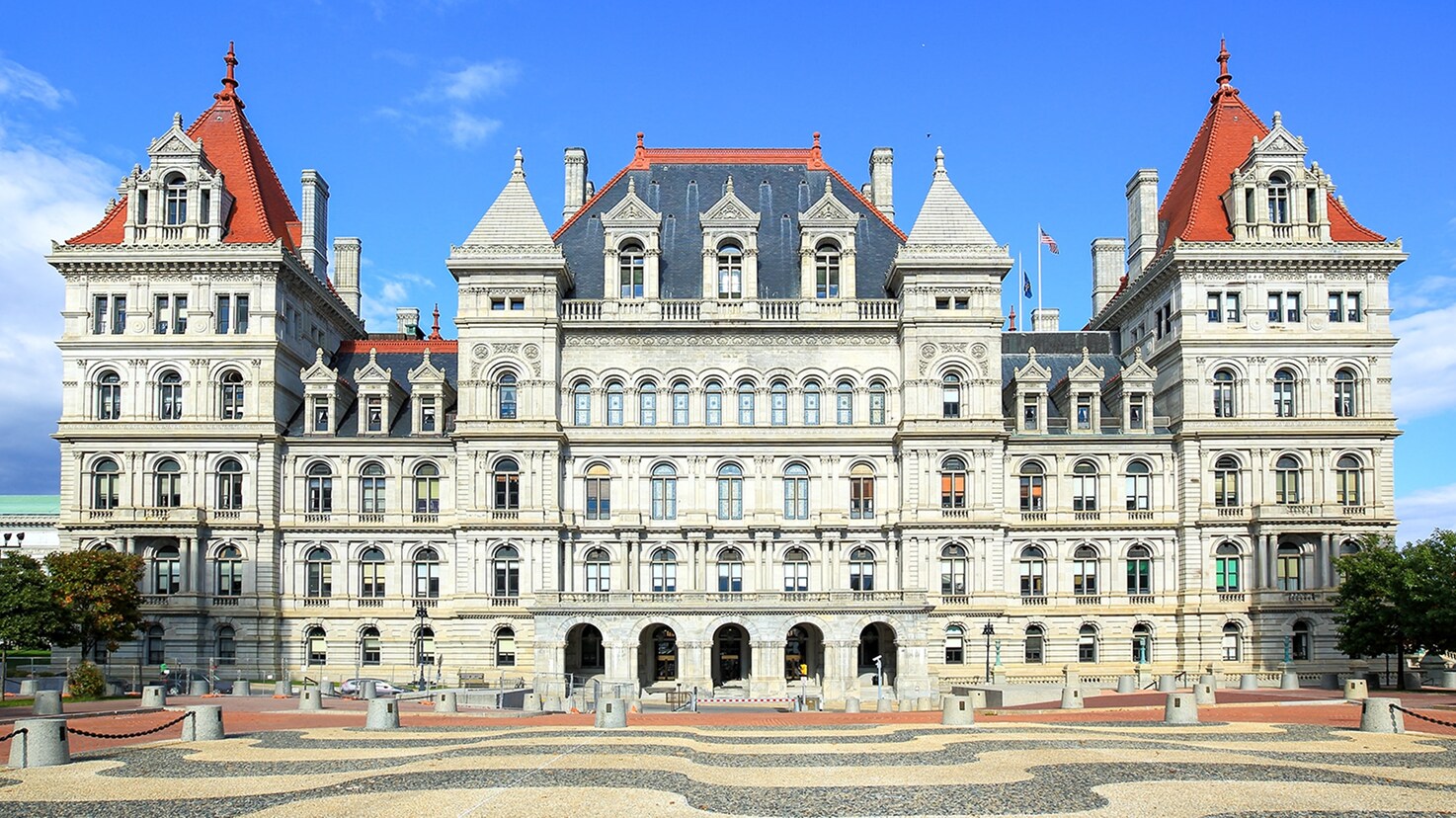 Exterior facade of the New York State Capitol building in Albany, New York, featuring the historic stone structure, arched entrances, and prominent red slate roofs.