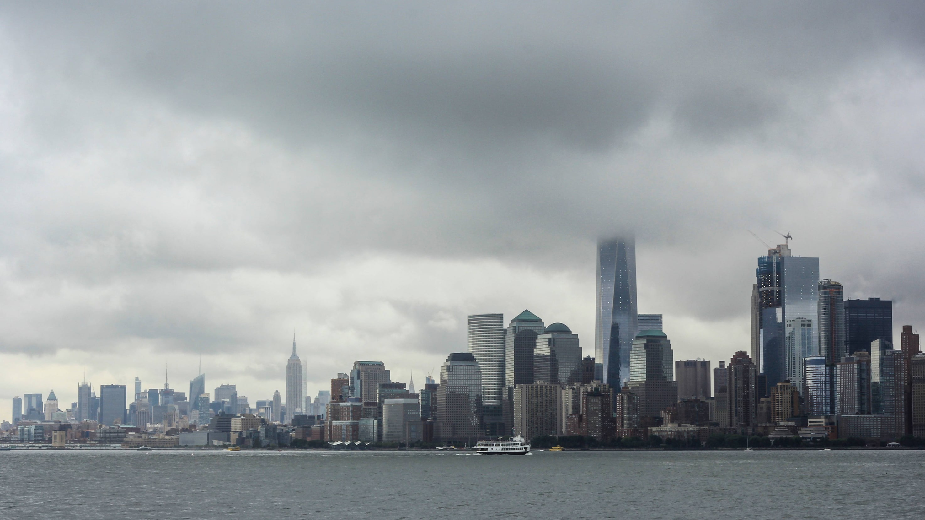 A cityscape with many tall buildings is shown under a cloudy sky, with a body of water and a few boats in the foreground